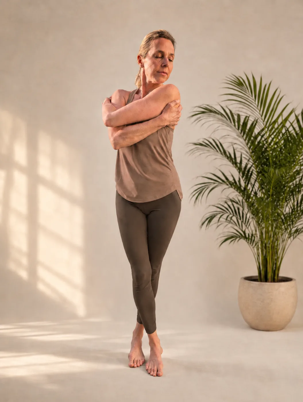 Woman with arms crossed in a gentle standing twist, eyes closed in warm afternoon light.