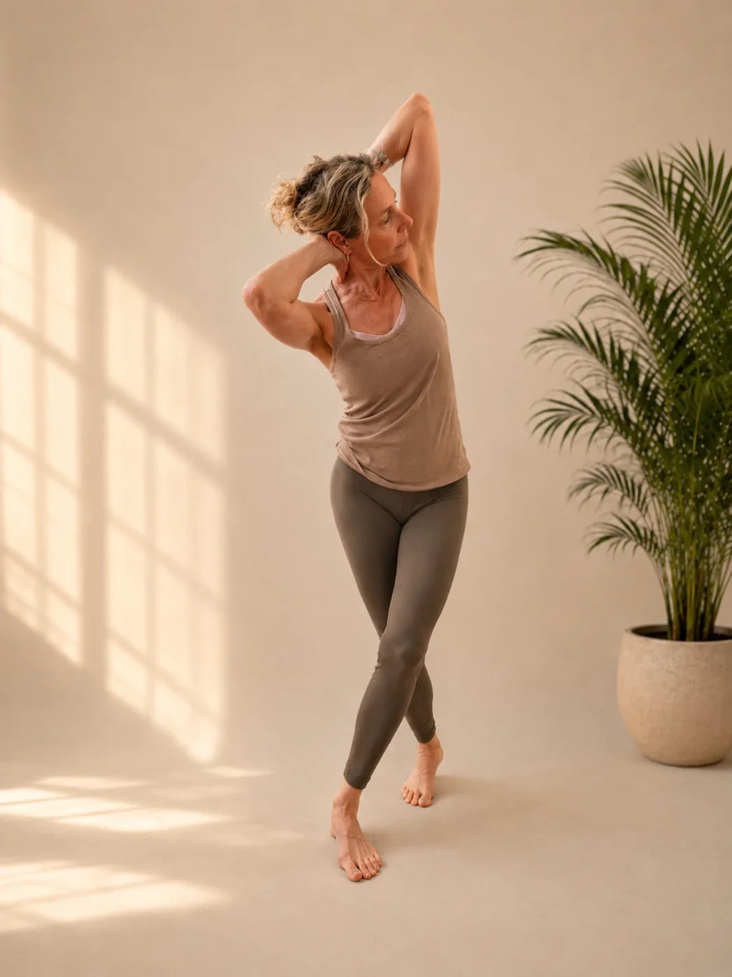 Woman in a standing spinal twist with both elbows open beside a potted palm.